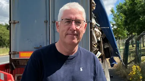Mark Hothersall stands by the back of the wagon. He has short white hair and glasses and is wearing a navy blue tshirt