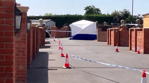 A residential street which has been blocked off by a police cordon. There is a white forensic tent at the top of the street.