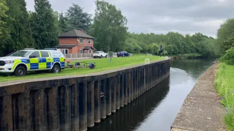 BBC/Phil Bodmer A lock next to water, surrounded by trees. On the bank is a police vehicle connected to an empty trailer. 