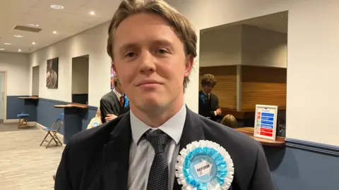 Grace Wood/BBC A man in a dark blue suit stands in hallway wearing a light blue and white rosette.