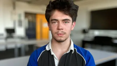 Martin Giles/BBC Jasper in a classroom. He has black hair and has a short beard and moustache. He is wearing a blue and black rugby shirt.