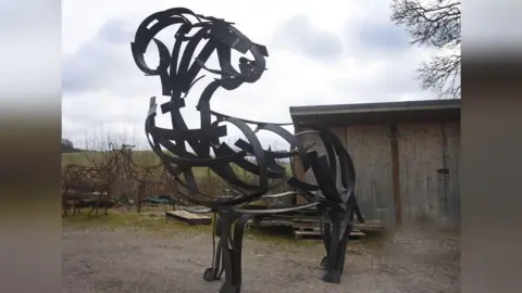 Andy Kay The 10ft (3m) high steel ram sculpture pictured in a yard by a shed. It is made of curved strips of dark metal, bent to form the outline of the body, head and horns of a ram. 