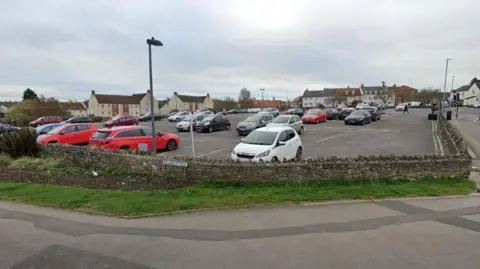 Rock Street Car Park in Thornbury. It is situated on a hill, on a bend in the road. It is surrounded by a brick wall and a pavement.