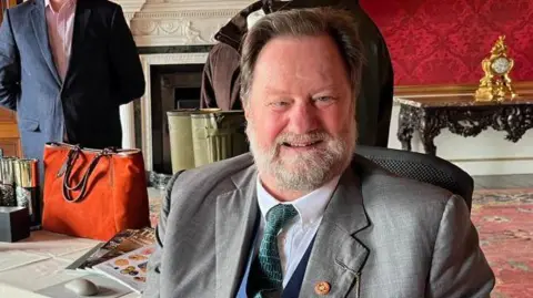 Champion News Service Lord Hartford, wearing a grey suit and a green tie in a room with red wall paper