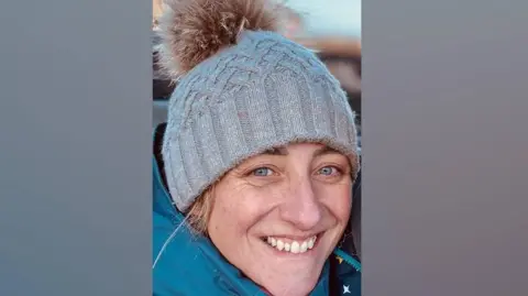Caroline Fancott-Beynon A close-up portrait of Caroline Fancott-Beynon. She is wearing a grey woolly hat with a furry light brown bobble and a bright blue jacket. She has blue eyes and blonde hair, and is smiling at the camera.