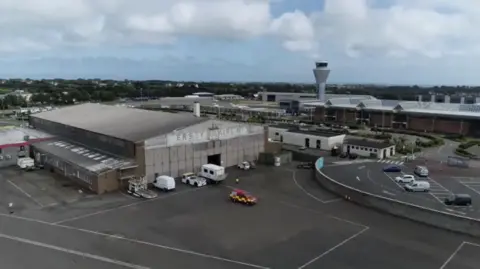 An aerial view of Jersey Airport, showing an old hangar with faded signage reading "Jersey Airlines Ltd" in the foreground. The structure appears weathered, with several vehicles, including a red and yellow emergency vehicle, parked nearby. In the background, the modern airport terminal and control tower are visible, along with surrounding infrastructure and greenery under a partly cloudy sky.