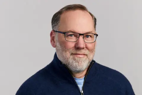 Reuters Fred Ramsdell, wearing a navy blue jumper and black-rimmed glasses, looks toward the camera in front of a neutral background.
