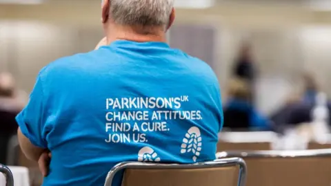 Parkinson's UK A man where's a Parkinson's UK blue top while sitting on a chair