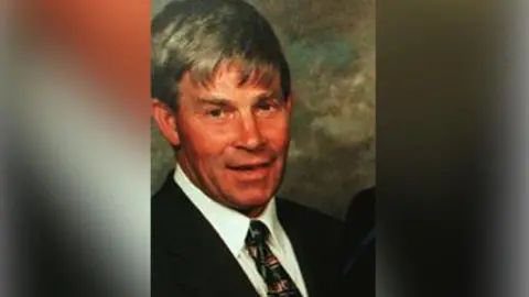 Bryan McCrory, a grey-haired man in a black suit, white shirt and green and red tie, partially smiles to the camera. Behind him is a dark grey/green background.