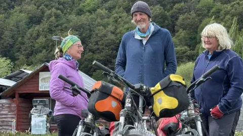 Pictured recently, Rona, Craig and Sophie standing next to their bikes, which are loaded with bags. There are mountains behind them.