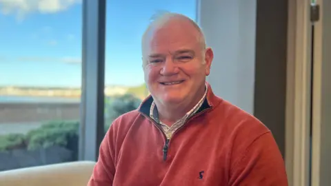 A man smiling wearing a red quarter zip sat in front of a glass window of the blurred sky.
