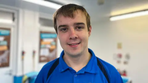 Euan Morrison has brown hair, brown eyes and is smiling at the camera. He is wearing a blue polo short with black stripes on shoulders.
