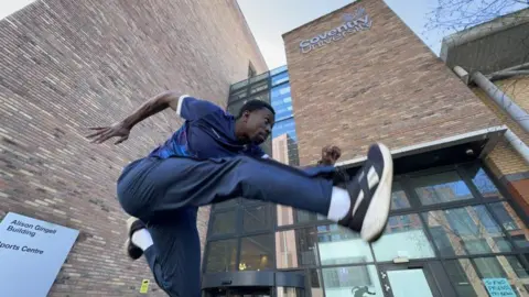 Coventry University Ethan is hurdling, picture mid-air in front of a university building 