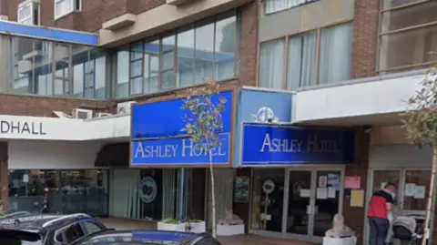 The blue entrance sign of the Ashley Hotel next door to a Costa and an M&S foodhall.