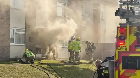Several firefighters on a grass area outside a low-rise flat block where smoke is billowing out from the bottom edge of the building.