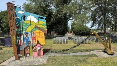 The colourful sculpture is captured in full in the park. Part of a gold anchor is attached with a chain to a sculpted bow of a ship which has designs, images and transcriptions on.