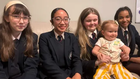 Four teenage schoolgirls and a young girl, aged about two - the schoolgirls are wearing dark school uniforms, with navy blazers and navy ties with a red stripe. The young girls has a yellow striped bottoms and a yellow vest. She has short blond hair set into short pigtails. 