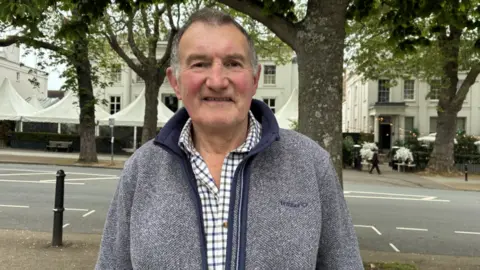 A man with a grey jacket and a check shirt underneath. He has grey hair. He's standing in front of a road with trees, white Regency-era buildings and white domed marquees behind him. 