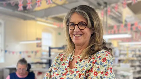 Emma Bridgewater - a woman wearing a patterned dress, glasses and a pearl necklace - is standing with her arms folded. She is smiling for the camera. Behind her is a factory with union jack bunting.