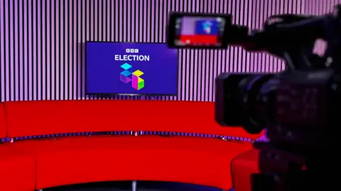 A red sofa in a broadcast studio, with wooden-panelled walls. There is a TV screen with a logo saying 'BBC Election 26' on it, on a purple background with white writing. In the foreground, a TV camera is visible.