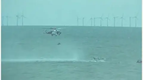 Michael Funnell A helicopter hovering over the sea and winching someone up. There are wind turbines in the background.