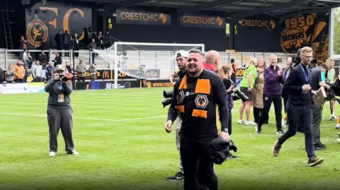 Dan McNamara with a Wolves scarf around his shoulders smiling as he stands on the pitch, with photographer sin the background