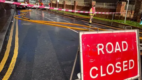 A road with a red road closed sign to the front of it, with a number of fire engine hoses scattered along the road. 