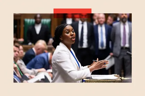 EPA Kemi Badenoch, viewed in profile, addresses the House of Commons from the despatch box. She wears a white jacket, a blue top and is holding a pen. The blurred outlines of other MPs are visible in the background.