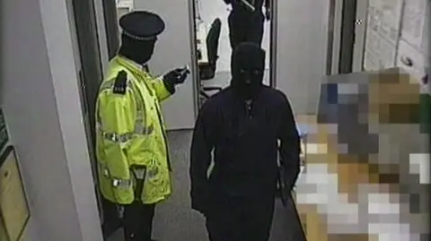 A gang member dressed as a police officer wearing a hi-vis jacket is standing with a gun in his hand. Two others, who are masked and wearing black are walking past him. There are notices on the wall in the depot.