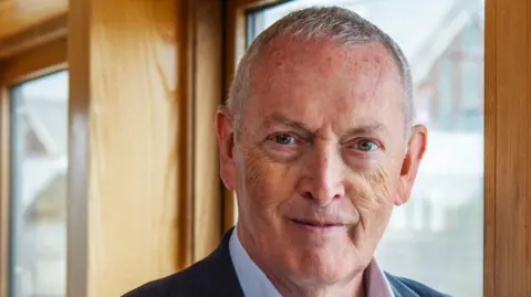 Lord Hutton, looking at the camera, smiling. He has short grey hair, and is standing by a window with a wooden frame. 