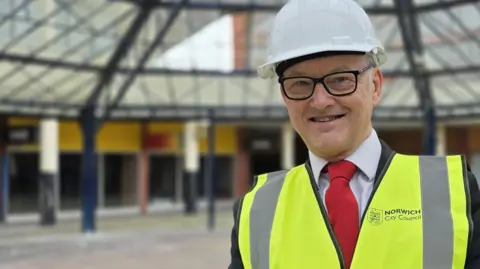 Mike Stonard is wearing a high-vis jacket over a suit with a red tie. He has a white hard hat on his head and is standing beneath a glass canopy.