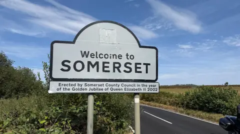 White road sign that says "Welcome to Somerset". There's a concrete road in the background and hedges and a field in the background. Blue skies and clouds in the background.