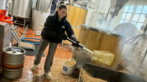 Hook Norton Brewery Georgina is scooping up an ingredient (perhaps barley) from a large container into a vat.