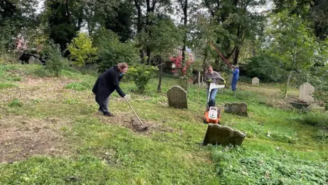 Dave Stewart Two female volunteers rake and mow the grounds of the graveyard. Several gravestones are dotted about the site and tall trees are visible in the background.