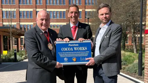 York Civic Trust The Lord Mayor of York, Sherriff's Consort and Dr Duncan Marks from the York Civic Trust, all wearing suits, hold the blue plaque. The factory is in the background.