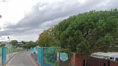 Google Maps A generic photo showing open blue gates, with a sign to the right of them with the letters CHCS on it. A building and grass are on the far left and trees are on the right of the image. 