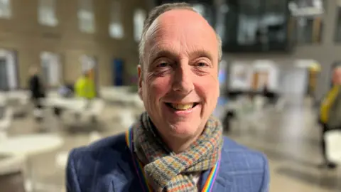 A middle-aged man smiles at the camera whilst wearing a tartan scarf and a rainbow lanyard with tables and other people obscured in the background.
