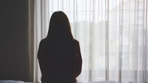 Getty Images A woman, who is silhouetted, sits on a bed staring out of a window