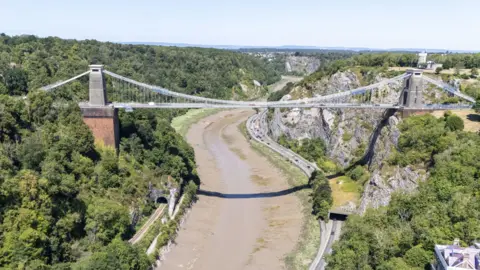 The Clifton Suspension Bridge in Bristol, a historic and iconic Victorian bridge that goes over the Avon Gorge in Bristol. The photo has been taken on a sunny day and cars are driving on the road beneath.