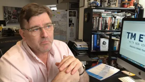 David McKelvey leaning on a desk looking pensive. He has brown hair and is wearing glasses and a pink shirt. He is surrounded by books, a computer, documents and maps.