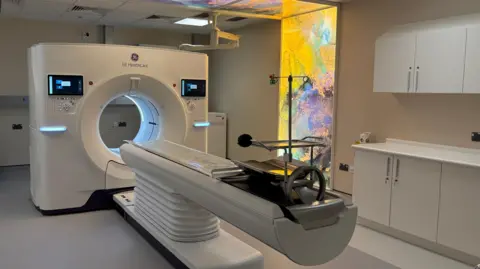 A CT scanner: a large square unit with a hole in the middle and a length of plastic, in the shape of a stretcher, sticking out into the room. The room is otherwise empty, aside from a series of kitchen-type units on the right-hand wall and a colourful light installation on the wall to the right of the scanner.