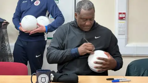 A man in a black sports jacket signs a white football with a permanent marker pen. There is a club branded mug on the table in front of him and another man, standing holding two footballs behind him.