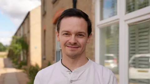 Dawid Wojtowicz/BBC Chris Tuczemskyi, standing in a shaded part of a street. He is smiling and wearing a white, collarless shirt. 