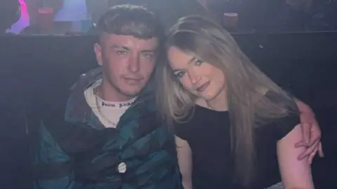 Libby Wolford Libby sat with her boyfriend at The Banc night club. Both are smiling looking at the camera. Her boyfriend is wearing a blue jacket and a white Palm Angels t shirt. Libby is wearing a black off the shoulder top.