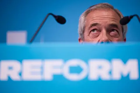 AFP A close up photo shows the Reform logo in white text against a light blue background on the front of a lecturn with the top half of Nigel Farage's head behind it as he looks up and away from camera with two small microphones in shot, as he delivers a speech at a press conference on 10 July in London.