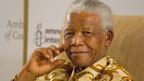 Head and shoulders of Nelson Mandela sitting in a gold fabric chair with his right hand touching his cheek as he smiles. He has white hair and is wearing a cream and gold shirt with a bold swirl pattern