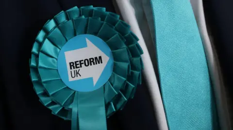 A close up of a turquoise coloured rosette with the words reform UK written in the centre. The rosette is pinned to the lapel of a black suit jacket being worn with a white shirt and turquoise tie.