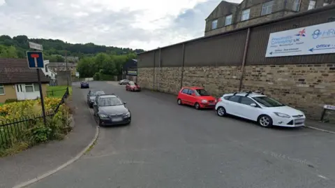 Google A residential/industrial street lined with parked cars. On one side appears to be a business premises, with homes on the other. Leafy trees are in the far background.