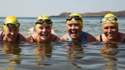 Four women pictured smiling in the sea wearing yellow swim caps and goggles on their head. Their legs are floating on the water's surface. 