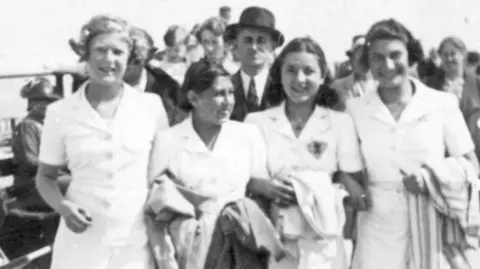 BBC/Jane Fellner A black and white photo showing four young women walking arm and arm along a seaside promenade wearing matching short-sleeved white shirts and shorts. Ilse (second from left) is smiling and looking to one side. The other three girls are smiling directly ahead. A man in a hat and a suit is directly behind them, but it's not clear if he's with the group.